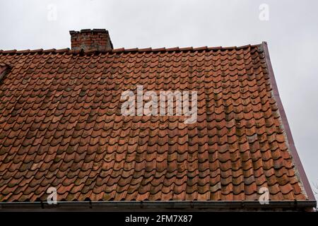 A fragment of an old shabby brown beige gable tiled roof of an old house with a brick chimney stack furnace with a rain gutter under it against the ba Stock Photo