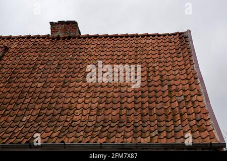 A fragment of an old shabby brown beige gable tiled roof of an old house with a brick chimney stack furnace with a rain gutter under it against the ba Stock Photo