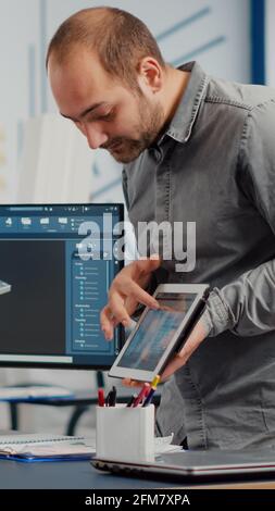 Industrial designer holding tablet discussing with woman engineer while working in CAD program, designing new components, comparing 3D prototype with project from notepad, using pc with two monitors Stock Photo