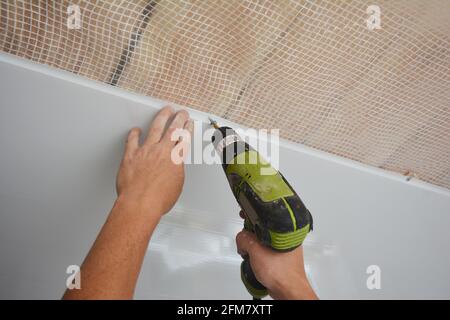 A builder is installing plastic ceiling panel, white PVC bathroom ceiling cladding in a bathroom using an electric screwdriver to attach it to the pla Stock Photo