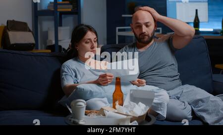 Young stressed married couple starting to cry sitting on couch holding papers reading bad news in legal document, got warning notice feel shocked looking frustrated. Eviction notification concept Stock Photo