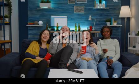Multiethnic friends laughing while watching comedy movie enjoying time spend together. Excited mixed group of people eating snacks, drinking beer having fun late at night during relaxed party Stock Photo