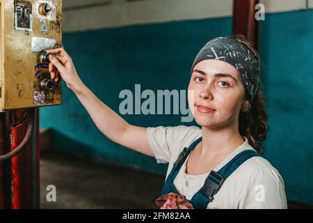 Gender equality. Portrait of a young woman in uniform working in a workshop or factory. Presses a button on the remote control. Stock Photo