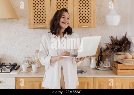 A smiling young woman in a kitchen, enjoying a video conversation, getting acquainted on a website. A woman spends time on a social network Stock Photo
