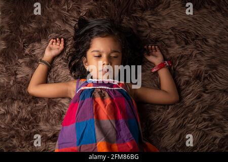Sleeping kid, a girl child showing different gestaurs who is lying on a synthatic rug which is very soft and brown in colour Stock Photo