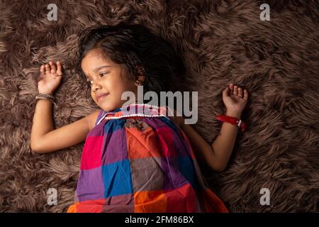 Sleeping kid, a girl child showing different gestaurs who is lying on a synthatic rug which is very soft and brown in colour Stock Photo