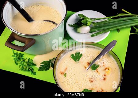 Dense creamy cauliflower soup in plate and pot, spilled soup on green kitchen board and parsley leaf, small plate with green and white onion. Stock Photo