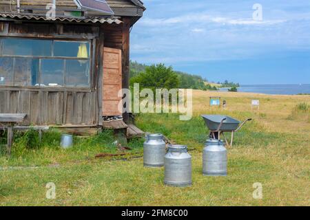 Old wooden house near Lake Baikal with milk cans and wheelbarrow on grass. The door is open. Clouds in the sky. Horizontal. Stock Photo