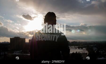 A rear view of a male security guard in uniform watching over the street from high floor. Stock Photo