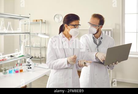 Diverse scientist in facial mask discuss test result using laptop at laboratory Stock Photo