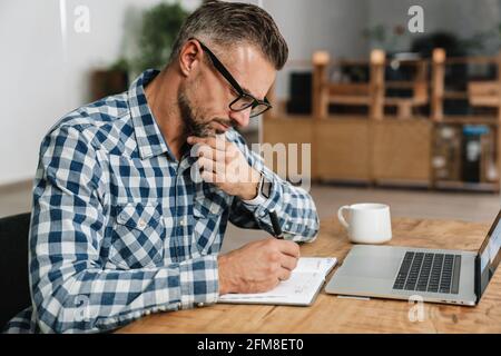 Focused grey man writing down notes while working with laptop in office Stock Photo
