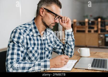 Focused grey man writing down notes while working with laptop in office Stock Photo