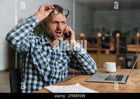 Shocked grey man talking on mobile phone while working with laptop in office Stock Photo
