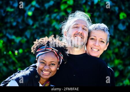 Mature caucasian couple and a young african woman posing hugging outdoor in a family pose in a garden with a leaves background. Stock Photo