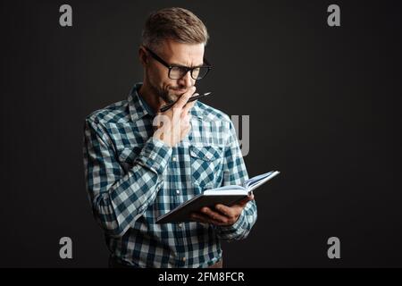 Photo of a thoughtful serious grey-haired man writing notes in notebook isolated over grey wall background Stock Photo