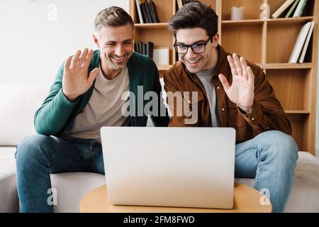 Happy young male gay couple using laptop computer for a video call while sitting on a couch at home, waving hands Stock Photo