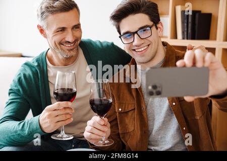 Happy gay couple hugging and drinking wine while taking selfie on cellphone at home Stock Photo