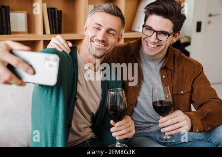 Happy gay couple hugging and drinking wine while taking selfie on cellphone at home Stock Photo
