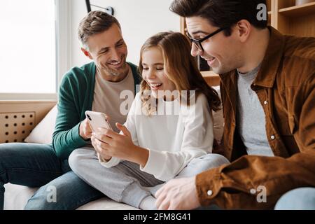 Happy gay family smiling and using mobile phone while sitting on couch at home Stock Photo
