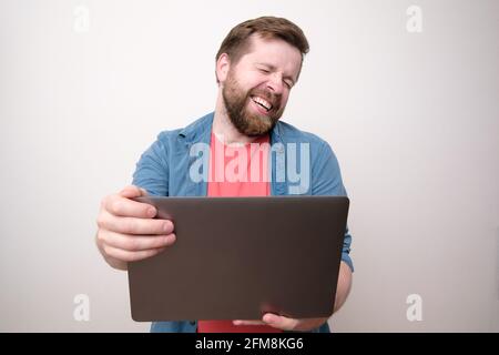 Cute man holds a laptop in hands and communicates via video communication, he laughs with eyes closed. Remote work or education.  Stock Photo