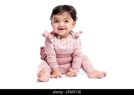 Cute female baby in lovely pink outfit smiling with mouth open. Attractive little baby sitting on floor and posing, isolated on white studio background. Concept of childhood. Stock Photo