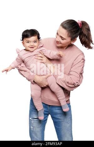 Portrait of cute mother holding daughter isolated on white studio background. Front view of young attractive woman hugging sweet adorable smiling baby girl while posing and looking at camera. Stock Photo