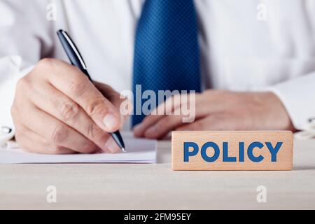 The word policy written on a wooden block with a businessman writing on a sheet of paper at the background. Insurance policy concept. Stock Photo