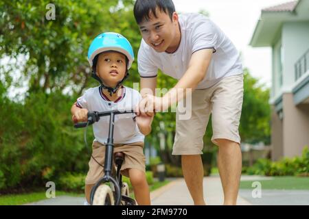 Asian father  teaching his child how to ride a bicycle in a neighborhood garden, fathers interact with their children throughout the day. Stock Photo