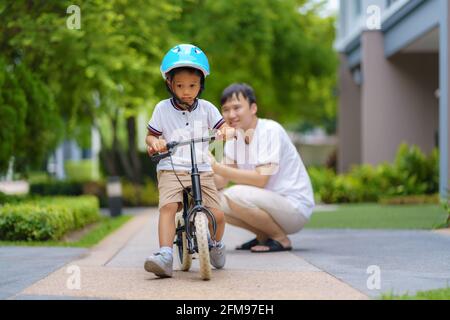 Asian father  teaching his child how to ride a bicycle in a neighborhood garden, fathers interact with their children throughout the day. Stock Photo