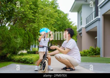Asian father wears a helmet for his son while teaching his child how to ride a bicycle in a neighborhood garden, fathers interact with their children Stock Photo