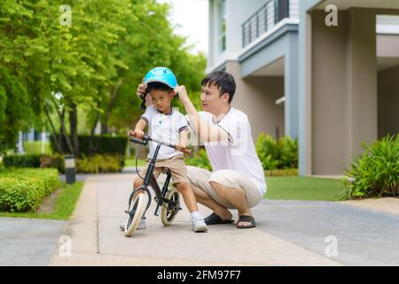 Asian father wears a helmet for his son while teaching his child how to ride a bicycle in a neighborhood garden, fathers interact with their children Stock Photo