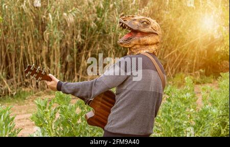 Man wearing dinosaur mask playing guitar outdoor in nature at sunset. Stock Photo