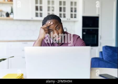 Worried African-American male freelancer using laptop for remote work from home, watching weary at the screen, holds forehead, has financial problem or project issues, unemployed man seeking for job Stock Photo