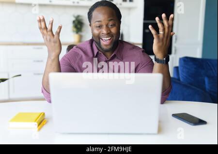 Surprised happy African-American male freelancer using laptop for remote work from home, amazed black man watching at the screen with mouth opened, raises hands, read good news, received job offer Stock Photo