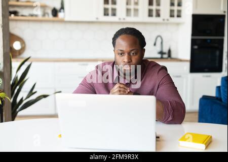 Concentrated African-American guy with dreadlocks staring at computer monitor sitting at the desk in home office, focused black man solving business tasks working with laptop remotely from home Stock Photo