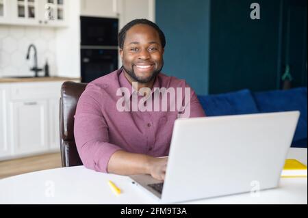 Cheerful African-American freelancer is using laptop for work on the distance, black guy sitting at the desk and typing, looks at the camera and laughs, enjoying remote work and free work schedule Stock Photo