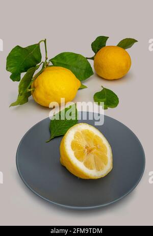 Still life of three lemons one of which is cut in half and placed on a gray plate with the light background. Stock Photo
