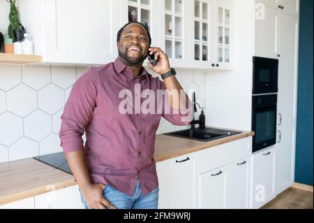 Hilarious black guy with dreadlocks in smart casual shirt enjoys phone conversation with friend or girlfriend, joyful African-American man laughs during pleasant phone conversation standing in kitchen Stock Photo