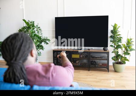 Back view African-American guy with dreadlocks holds remote controller and switching tv channels, watching TV sitting on the couch at home, the lcd TV with empty black screen, copy space, mockup Stock Photo