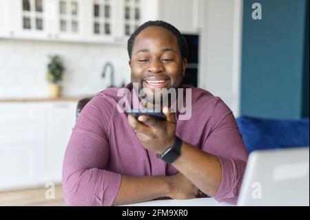 Cheerful African-American man with locks hair sending voice message sitting at home office, positive black guy in casual shirt using voice recognition app, recording audio report Stock Photo