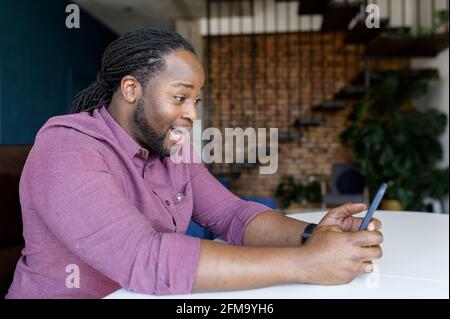 Amazed black guy surprised with unexpected good news sitting in modern apartment and staring at the smartphone screen, shocked happy African-American man received best deal by email on mobile phone Stock Photo