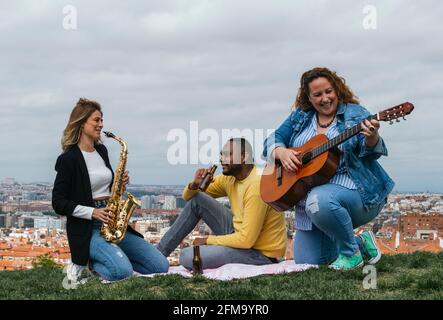 Happy interracial friends sitting in a park playing saxophone and guitar and laughing Stock Photo