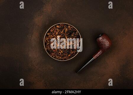 Close up round tin full of ready rubbed long coarse cut tobacco blend and handmade briar smoking pipe over grunge brown background, elevated top view, Stock Photo