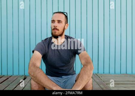 Bearded young man wearing sunglasses is confidently looking away at the Isla Canela beach with a blue wooden wall background Stock Photo