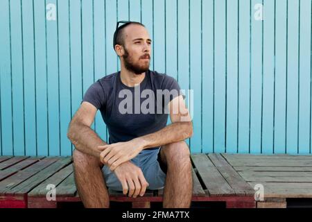 Bearded young man wearing sunglasses over the head is confidently looking away at the Isla Canela beach with a blue wooden wall background Stock Photo