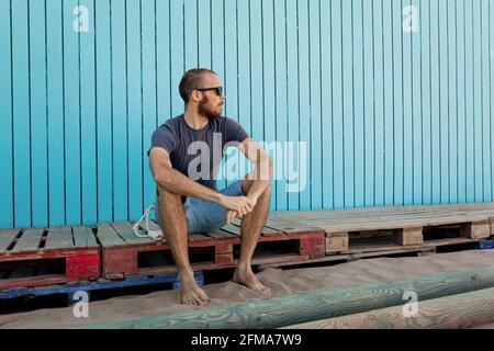 Bearded young man wearing sunglasses is confidently looking away at the Isla Canela beach with a blue wooden wall background Stock Photo