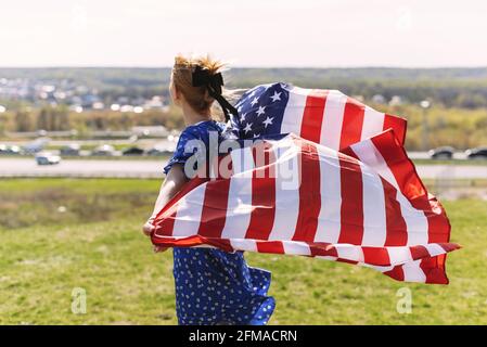 A young woman with the national flag of the USA on her shoulders against the backdrop of the city celebrates the Independence Day of the United States
