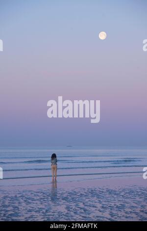 Solitary woman taking an early morning pre-dawn stroll on the beach at low tide, with the moon above. Brighton & Hove, Sussex, England, UK Stock Photo