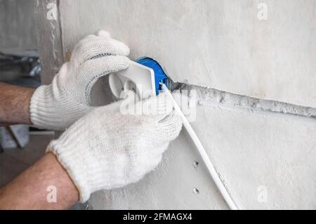 Close-up on the hands of a Caucasian male electrician installing a white AC outlet on a gray wall of a house.Home repairs during the pandemic and stay Stock Photo