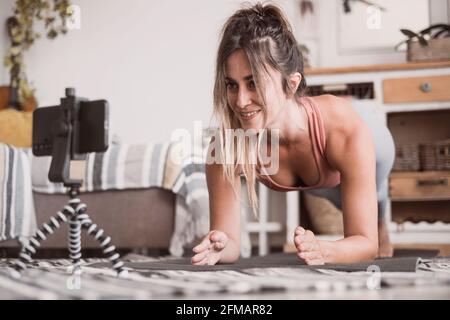 Young sporty woman working out at home, teenager doing fitness exercises on living room floor for buttocks body shaping using online personal training program with phone, doing yoga pilates indoors Stock Photo